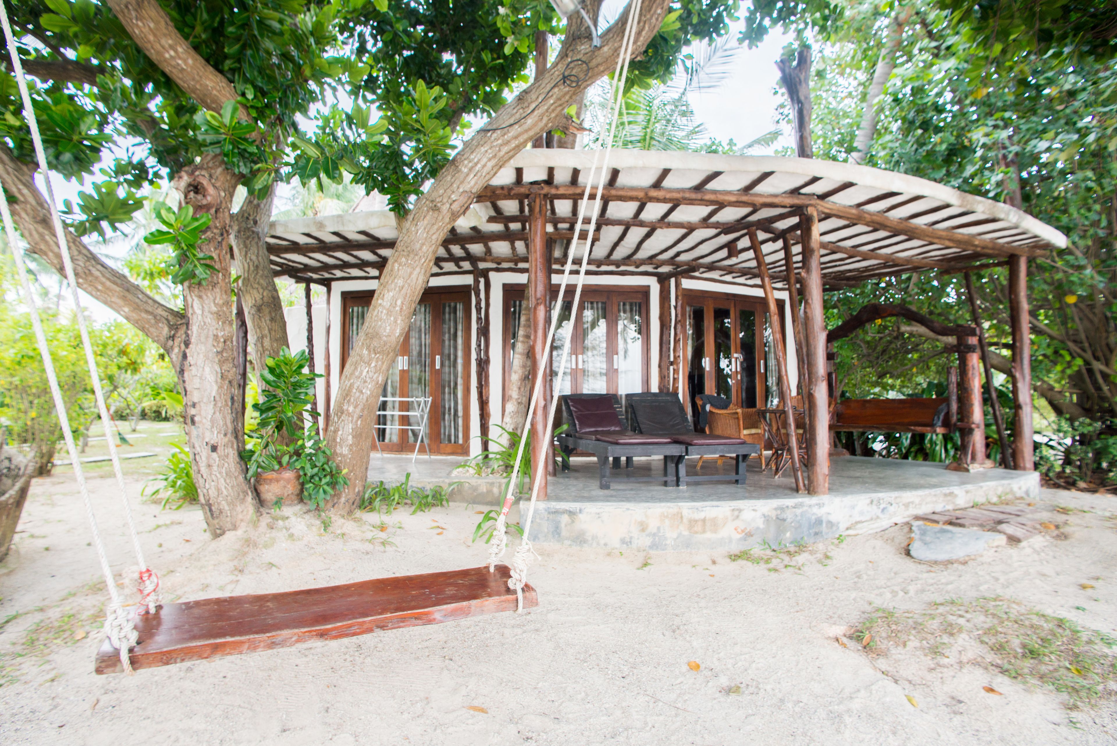 A wooden pavilion with a swing, surrounded by trees and a sandy ground.