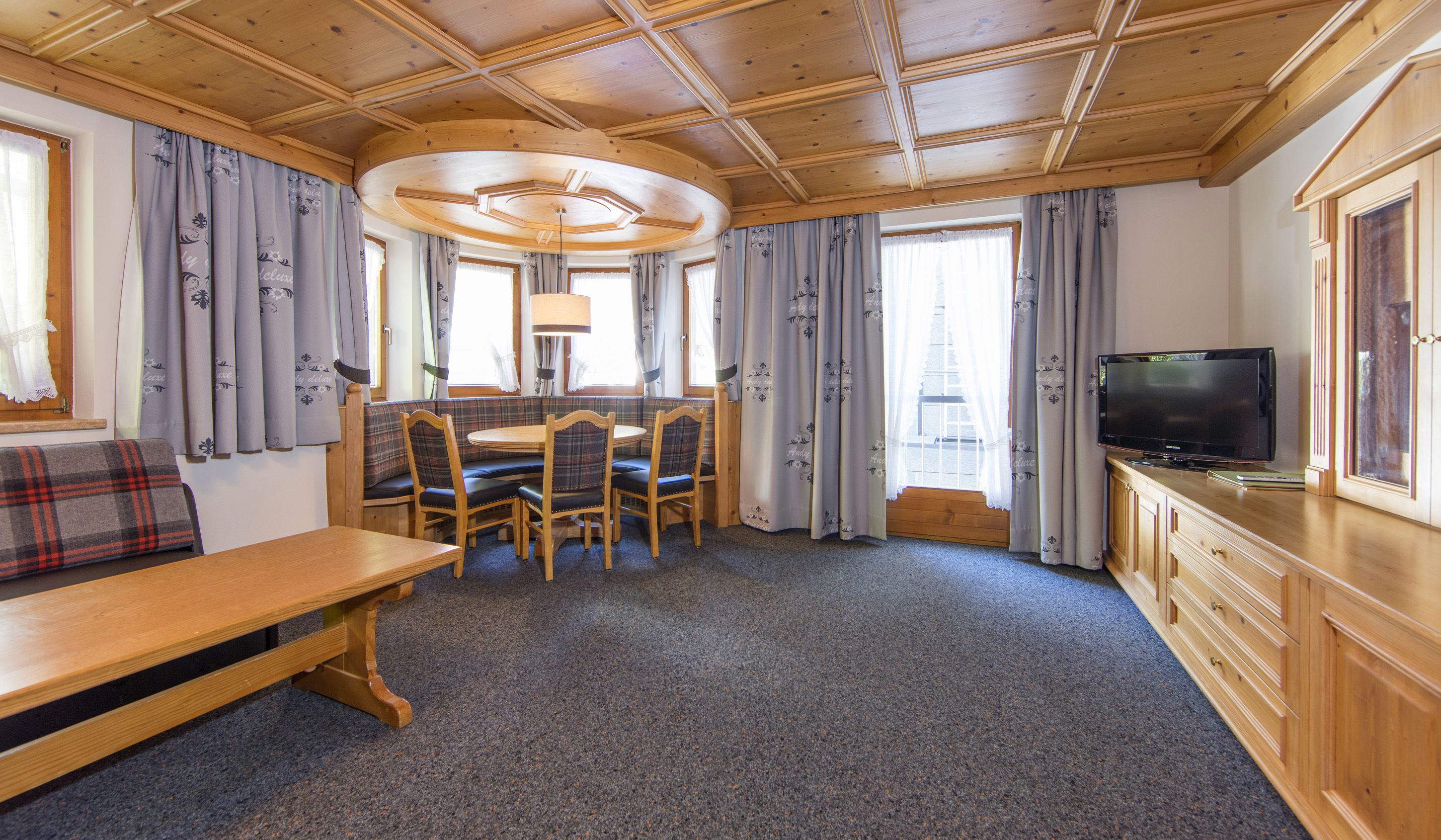 A cozy room with a wooden ceiling, a dining table with chairs, a bench, a television on a cabinet, and curtains on the windows.