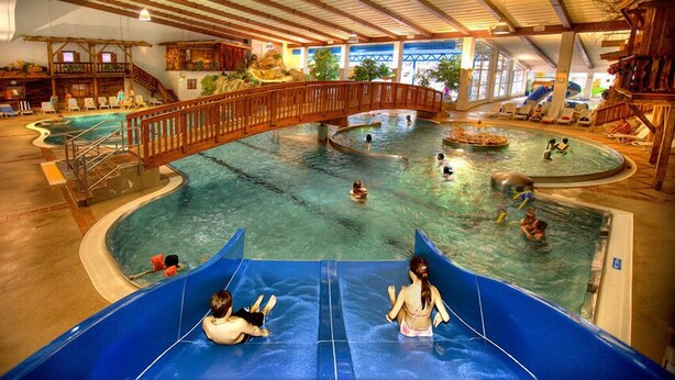 Un parco acquatico al coperto con una piscina centrale, scivoli e un ponte di legno.