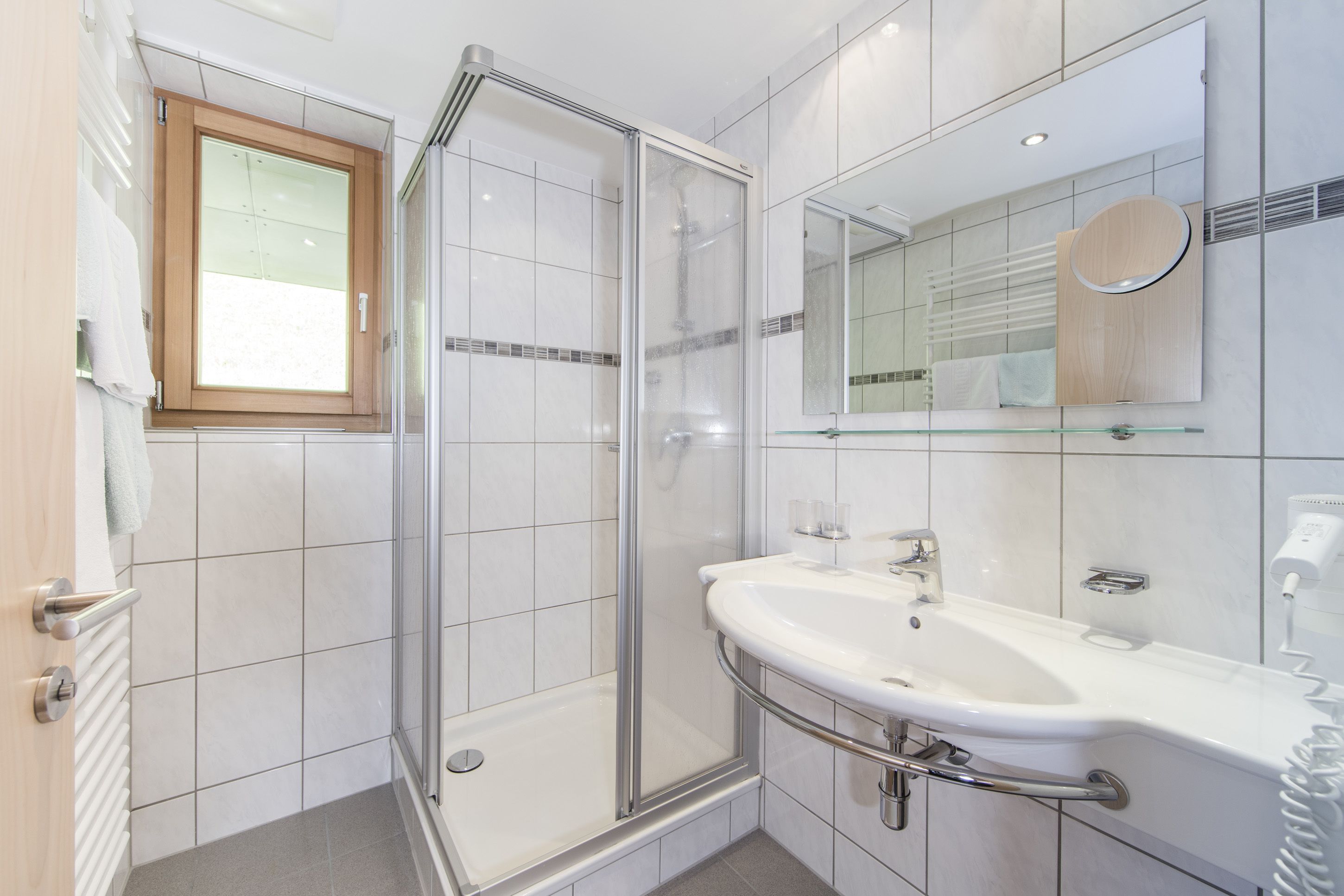 A modern bathroom with a glass shower enclosure, a white sink with a mirror above, and a wooden-framed window.