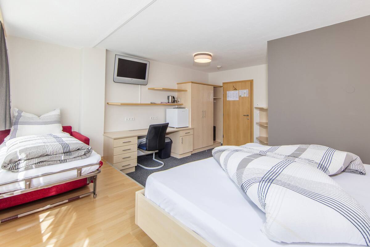 A bedroom with two beds, a desk, a chair, a wardrobe, a television, and a small refrigerator.