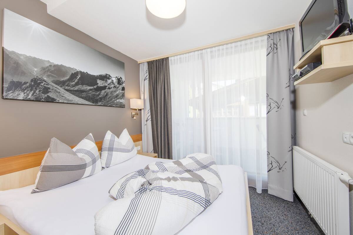 A hotel room with a bed, a mounted TV, a window with sheer curtains, and a wall-mounted artwork.