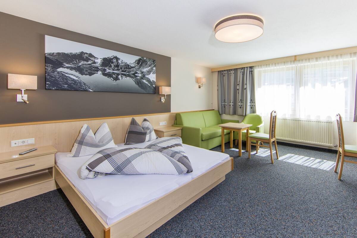 A hotel room with a bed, a green armchair, a wooden table, and a painting on the wall.