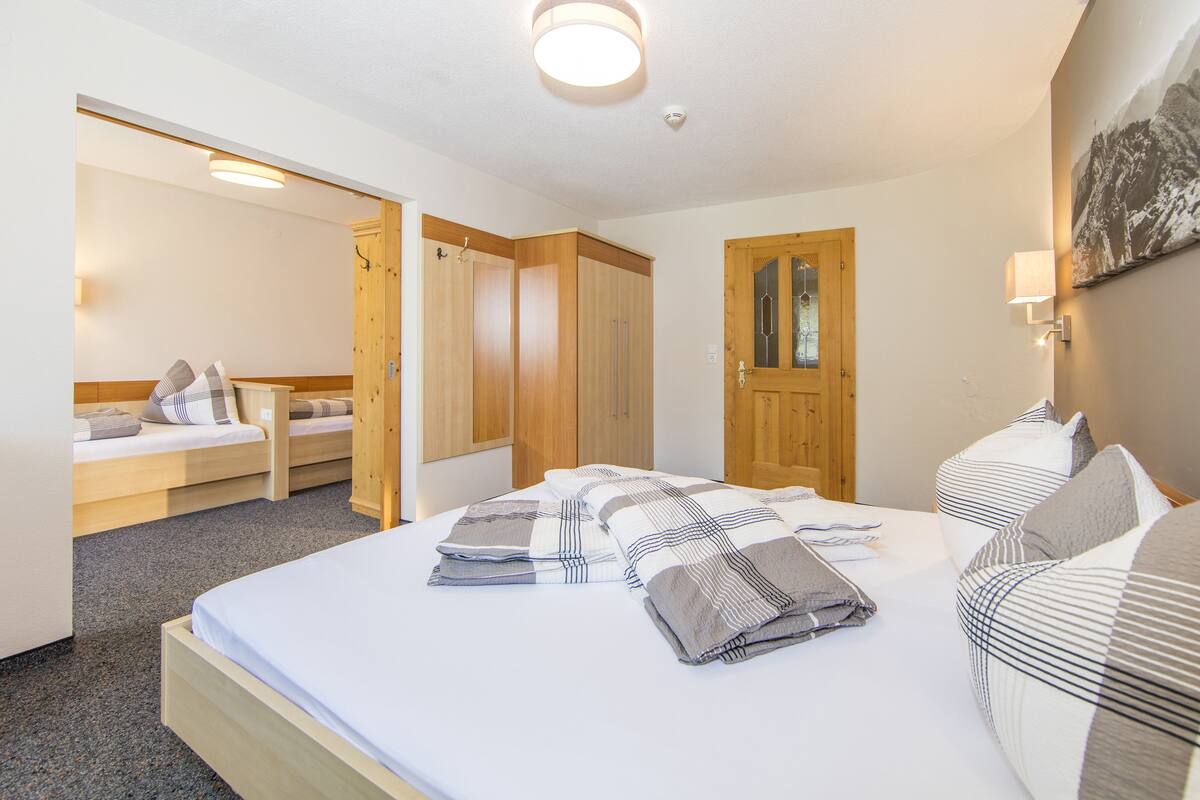 A hotel room with a large bed, a wooden wardrobe, a small seating area, and a door leading to another room.
