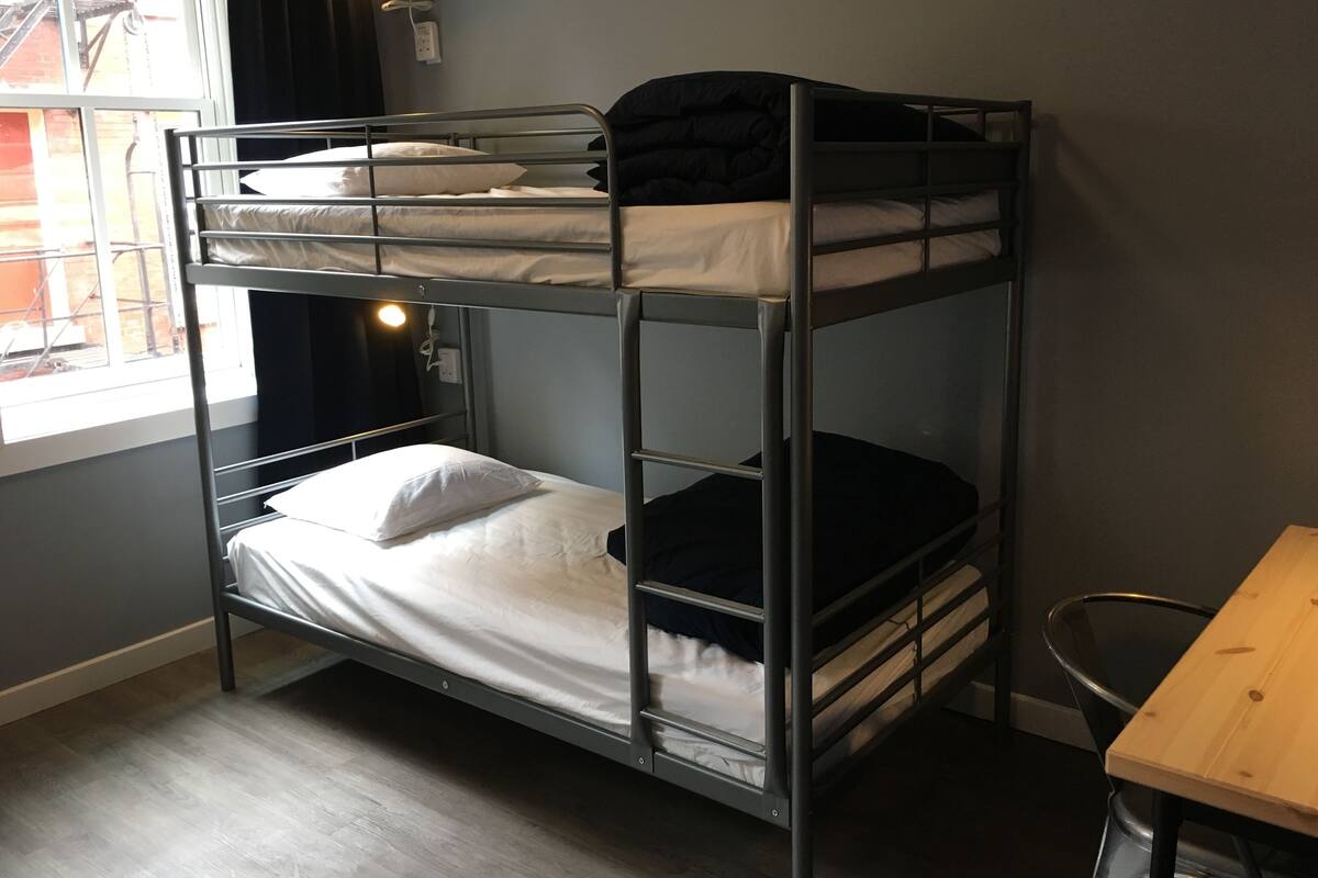 A bunk bed with a desk and chair, a window with curtains, and a wall socket.