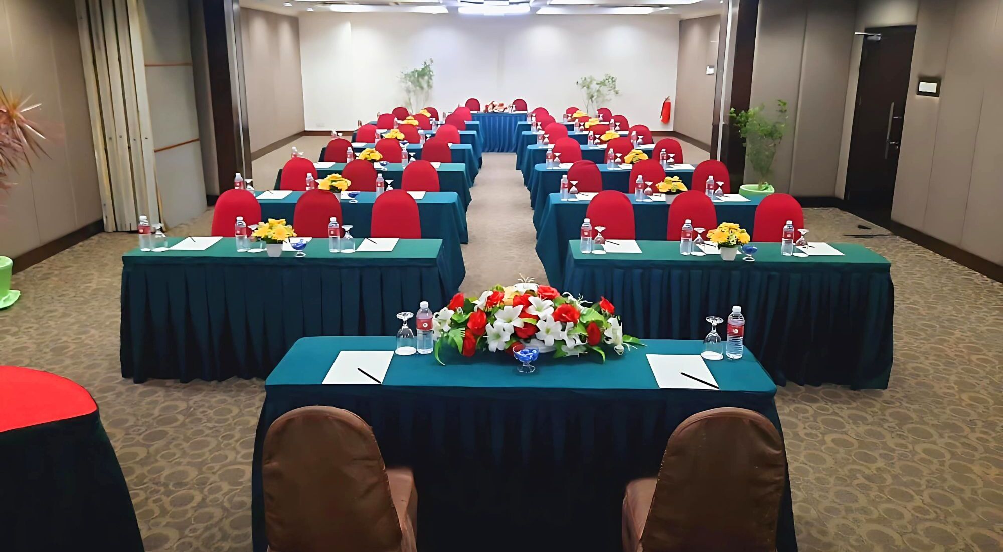 A conference room with rows of tables, each with red chairs, water bottles, and floral centerpieces.