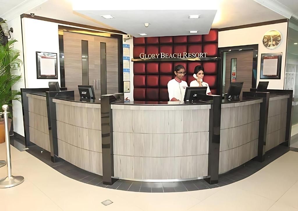 A reception desk at Glory Beach Resort with two staff members, a clock, and a framed picture on the wall.