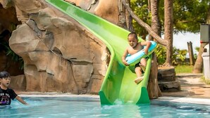 A child sliding down a green water slide into a pool with a rocky backdrop.