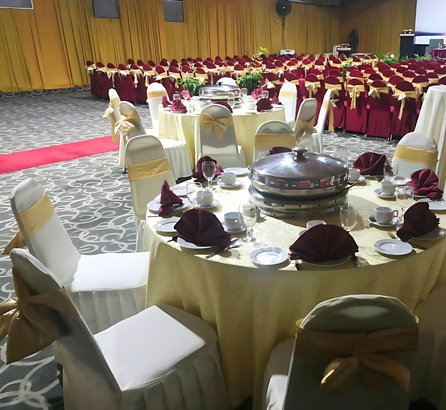 Un salón de banquetes con mesas redondas dispuestas para un evento formal, con sillas blancas adornadas con cintas doradas, manteles rojos y una alfombra roja que conduce al escenario.