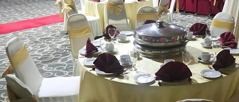 Un salón de banquetes con mesas redondas dispuestas para un evento formal, con sillas blancas adornadas con cintas doradas, manteles rojos y una alfombra roja que conduce al escenario.