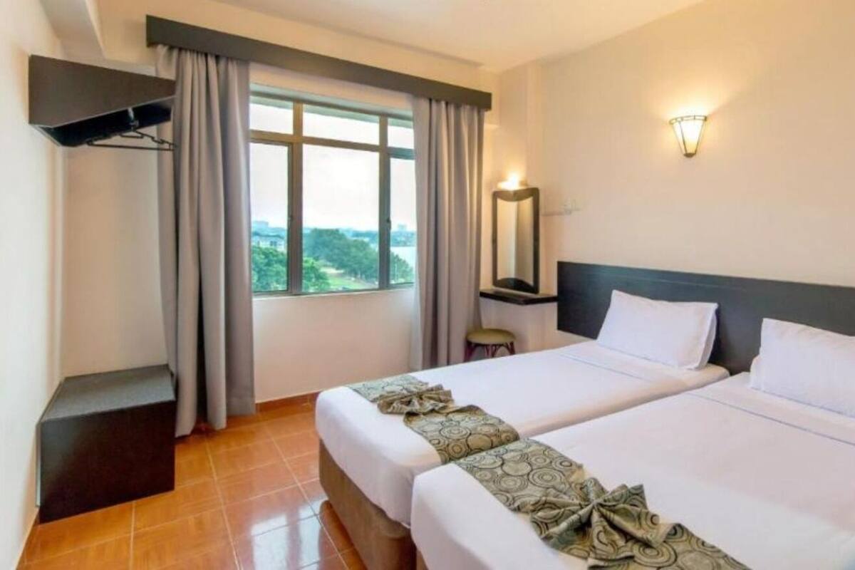 A hotel room with two beds, a large window with curtains, a bedside table, a mirror, and a wall-mounted light fixture.