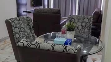 A dining area with a glass table and patterned chairs, a ceiling fan, and a view of a living room with a TV and curtains.