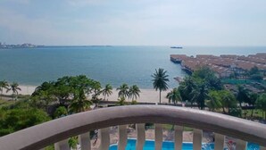 A coastal view from a balcony overlooking a beach, a pool, and a distant cityscape.