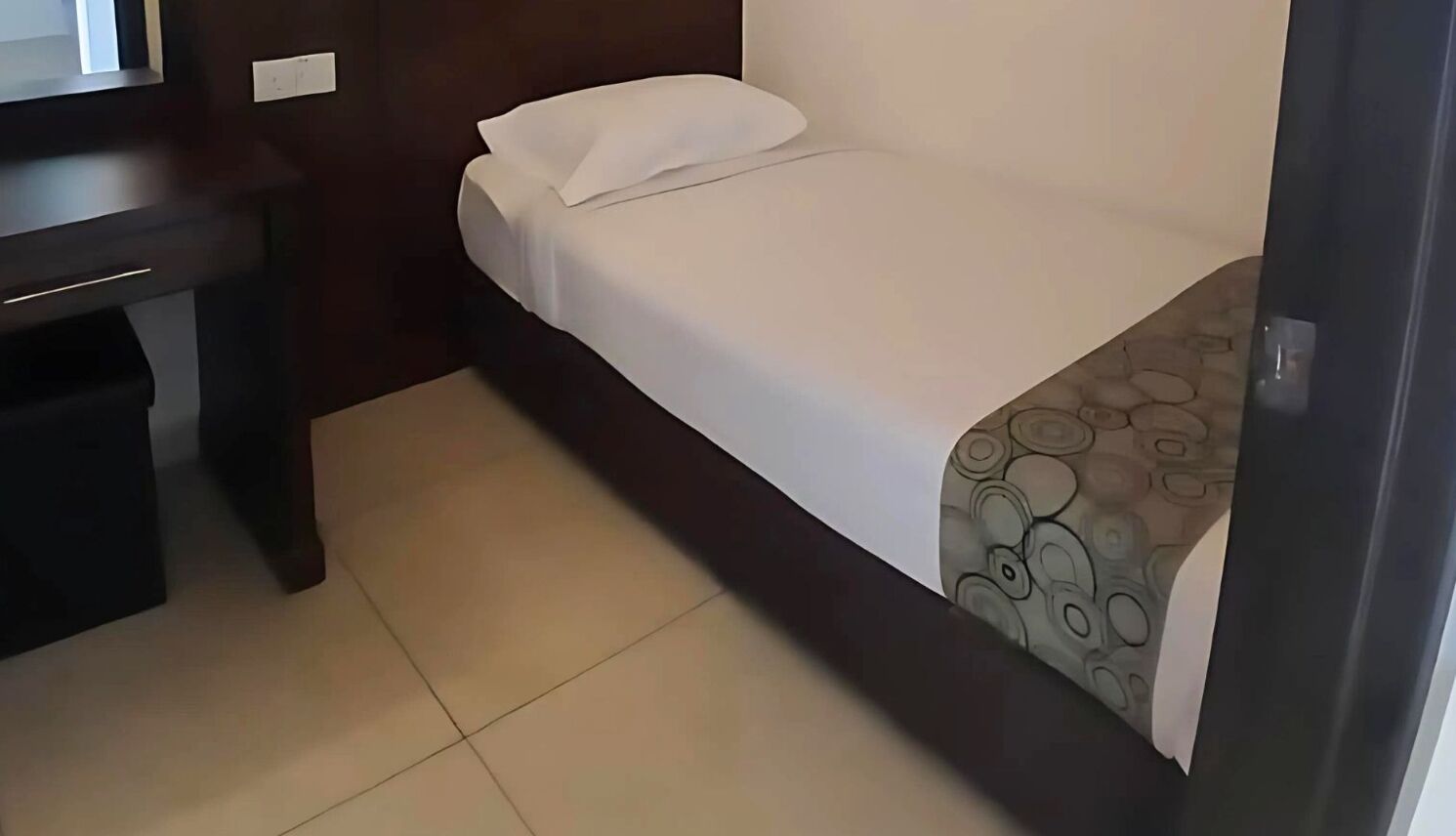 A small, neatly made bed with a white sheet and a patterned blanket, a dark wooden nightstand, and a tiled floor.