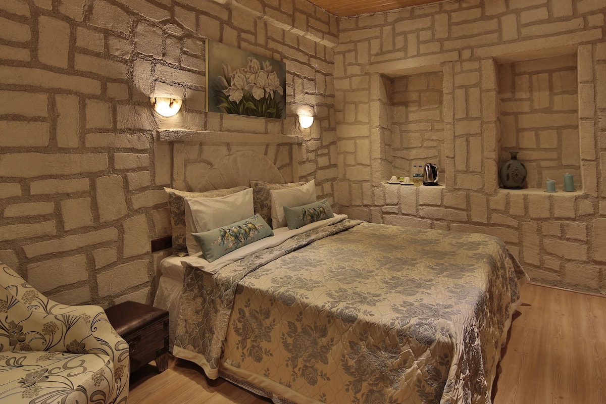 A bedroom with a stone wall, a bed with floral bedding, a nightstand, a chair, and a shelf with candles and a kettle.