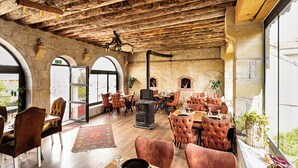 Interior rústico com paredes de pedra, vigas de madeira e portas em arco. Mesas e cadeiras dispostas para refeições.