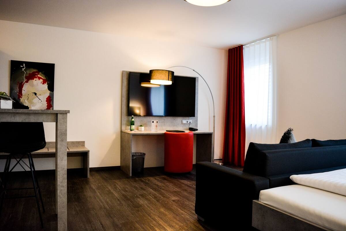 A modern hotel room with a wooden floor, a black sofa, a red ottoman, a small desk with a chair, a flat-screen TV mounted on the wall, and a window with red curtains.