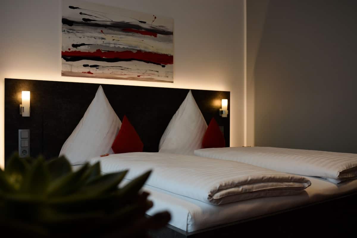 A hotel room with a bed, two bedside tables, a wall-mounted abstract painting, and a potted plant in the foreground.