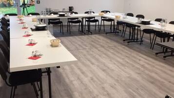 Sala de conferências com várias mesas, cadeiras e uma janela com vista para árvores.