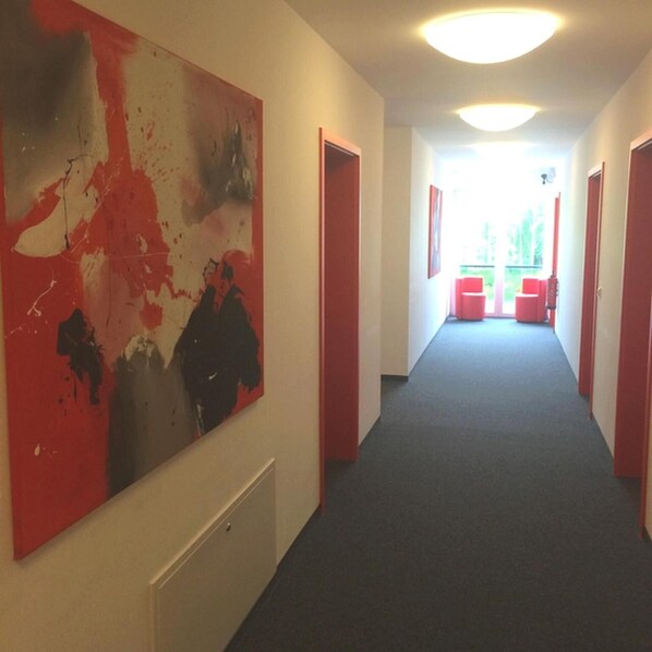 A hallway with a large abstract painting on the left, red doors, and a bright ceiling light. - Campushotel GmbH (Hagen)