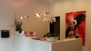 A reception desk with a white counter, a potted plant, and a red abstract painting on the wall. - Campushotel GmbH (Hagen)
