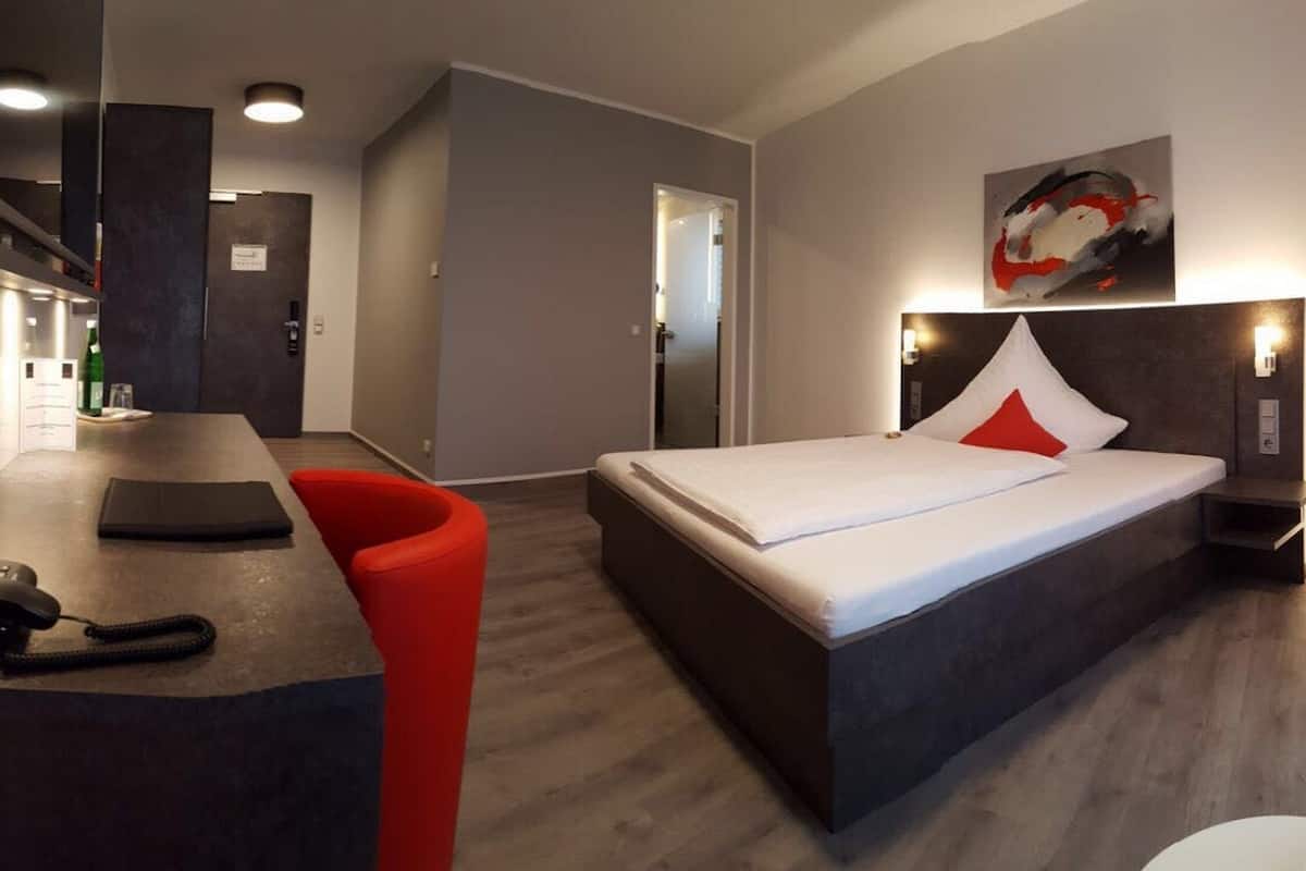 A modern hotel room with a large bed, a desk with a telephone, and a red chair.