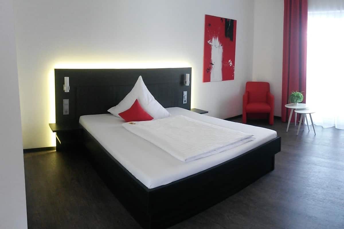 A modern hotel room with a large bed, a red chair, a small table with a plant, and a red and white abstract painting on the wall.