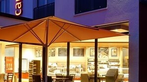 A restaurant with an outdoor seating area, a large umbrella, and a visible menu board. - Campushotel GmbH (Hagen)