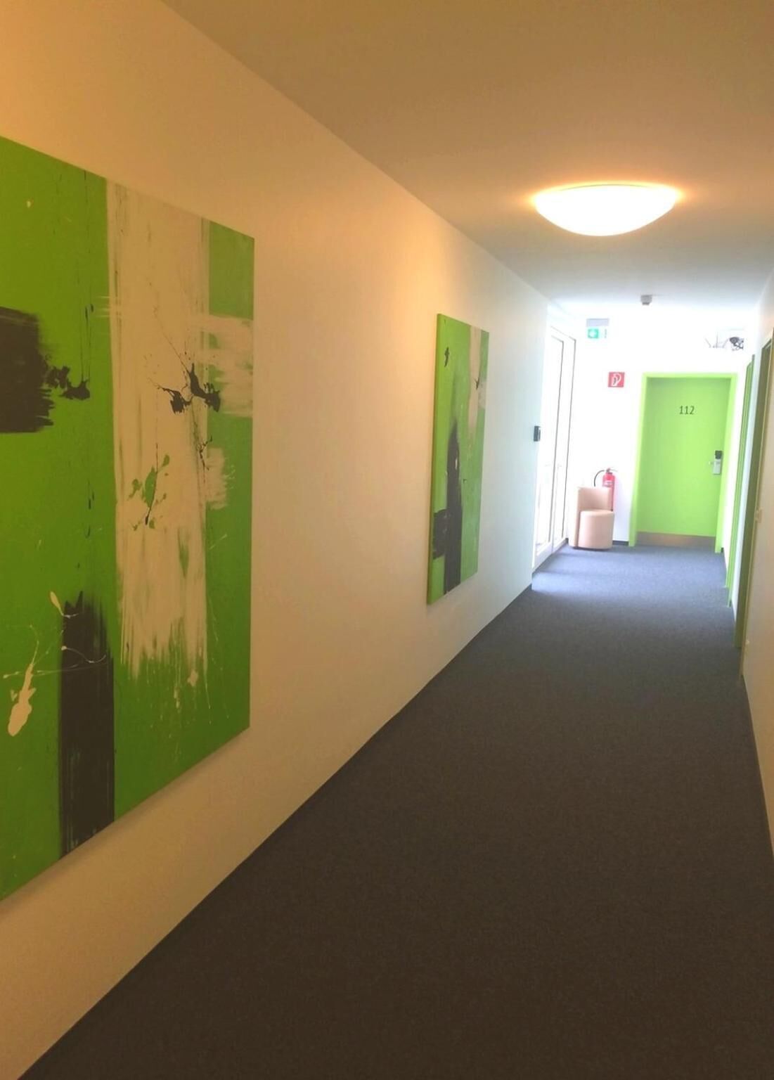 A hallway with green and white abstract paintings on the walls, a fire extinguisher, and a door with the number 112.
