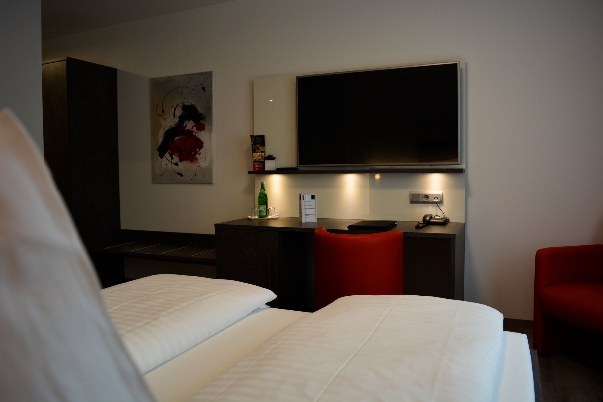 Ein Hotelzimmer mit Bett, Schreibtisch, Stuhl, Fernseher und einem Gemälde an der Wand.