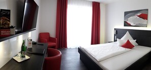 A hotel room with a bed, a desk, a chair, a TV, and a window with red curtains. - Campushotel GmbH (Hagen)