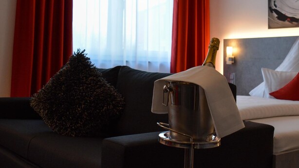 A hotel room with a bed, a black sofa, red curtains, and a bottle of champagne on a table. - Campushotel GmbH (Hagen)