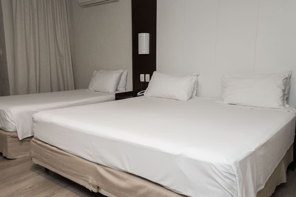 A hotel room with two single beds, a dark-colored wall, a white air conditioning unit, and a light-colored curtain.
