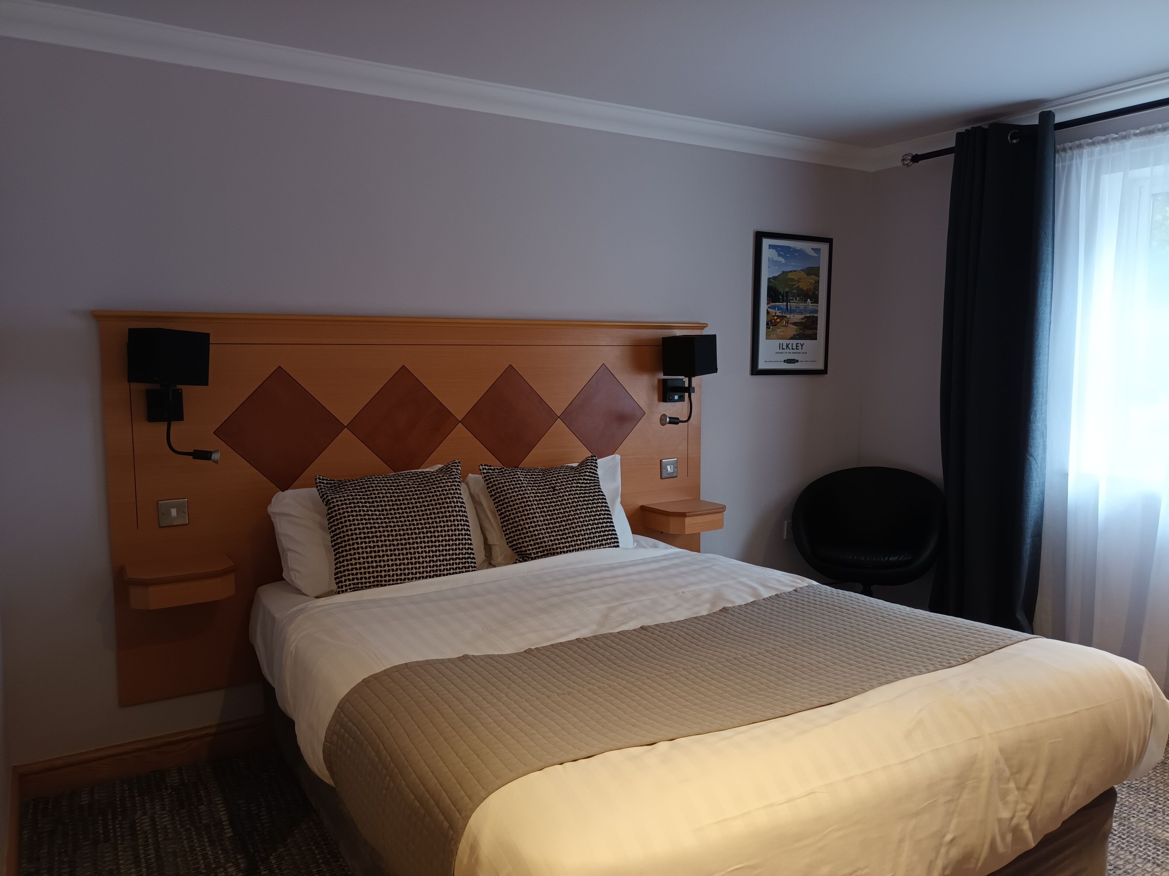 A hotel room with a bed, headboard, bedside tables, a wall-mounted TV, a framed picture, and a window with curtains.