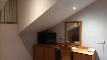Une petite pièce à la lumière tamisée, avec un plafond en pente, un bureau en bois sur lequel se trouve une télévision, une chaise et un miroir.