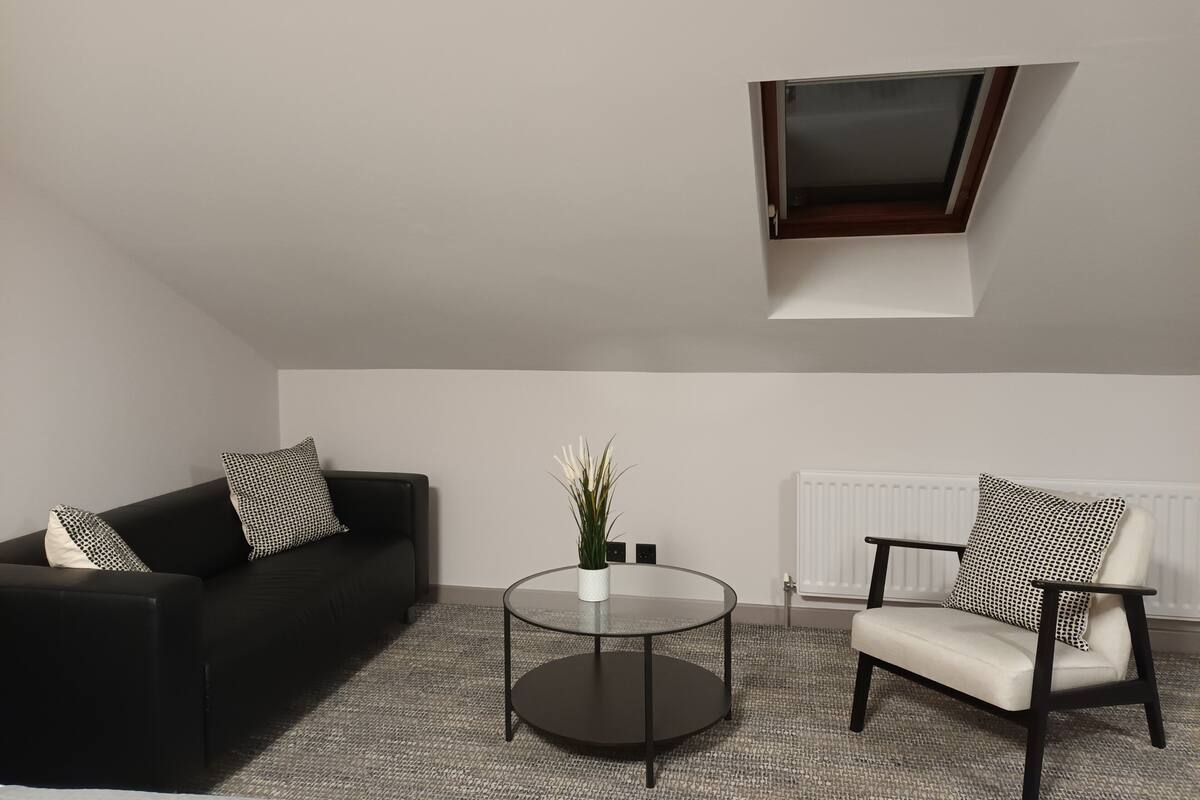 A modern living room with a black sofa, a white armchair, a glass coffee table, and a potted plant.