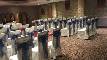 De longues rangées de chaises blanches, ornées de rubans bleus, dans une salle de banquet au sol recouvert d’un tapis à motifs et aux murs en briques.