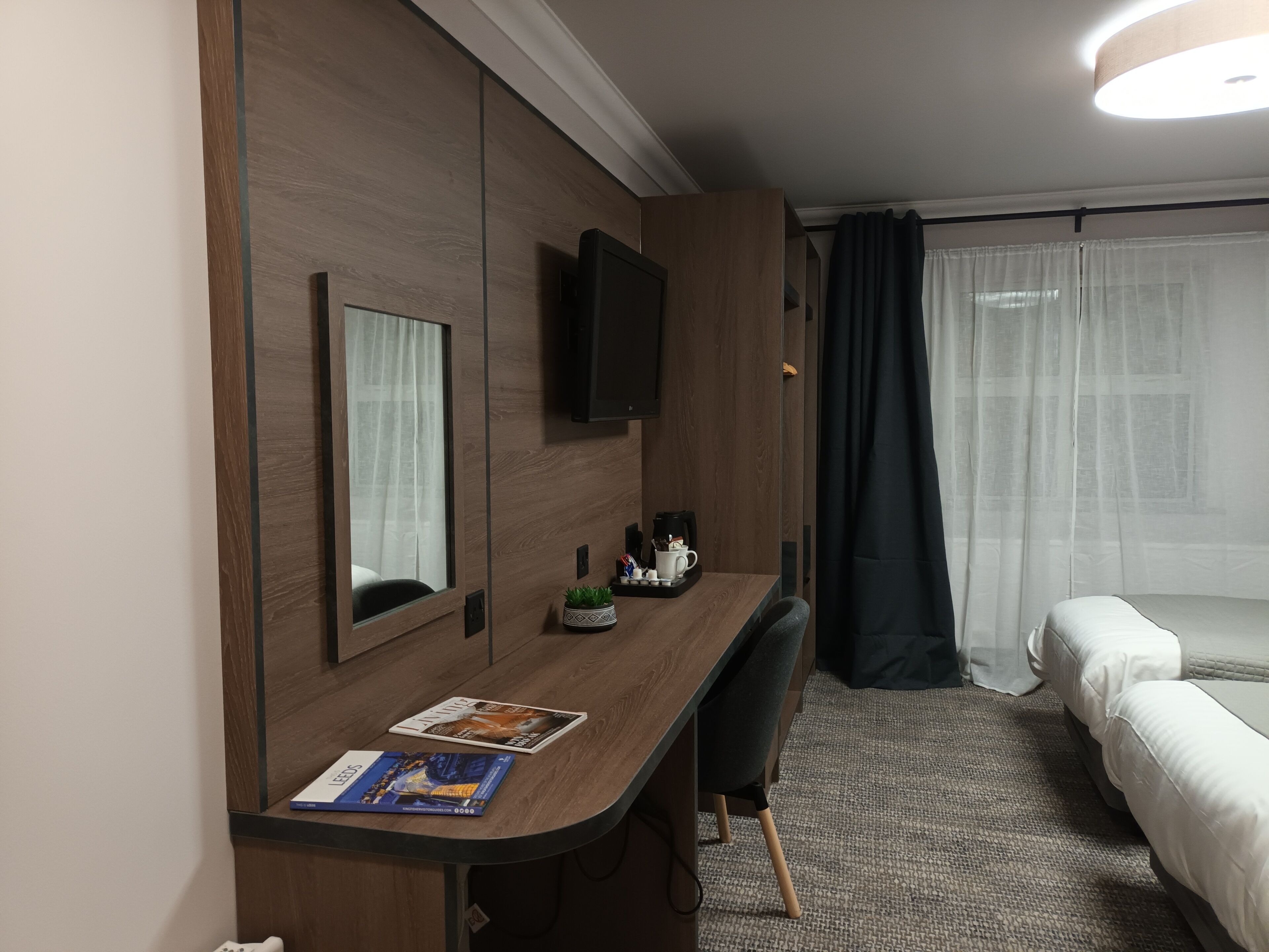 A hotel room with a wooden desk, a flat-screen TV, a bed, and a window with sheer curtains.