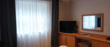 Quarto de hotel com cama, televisão sobre um móvel de madeira, espelho na parede e janela com cortinas.
