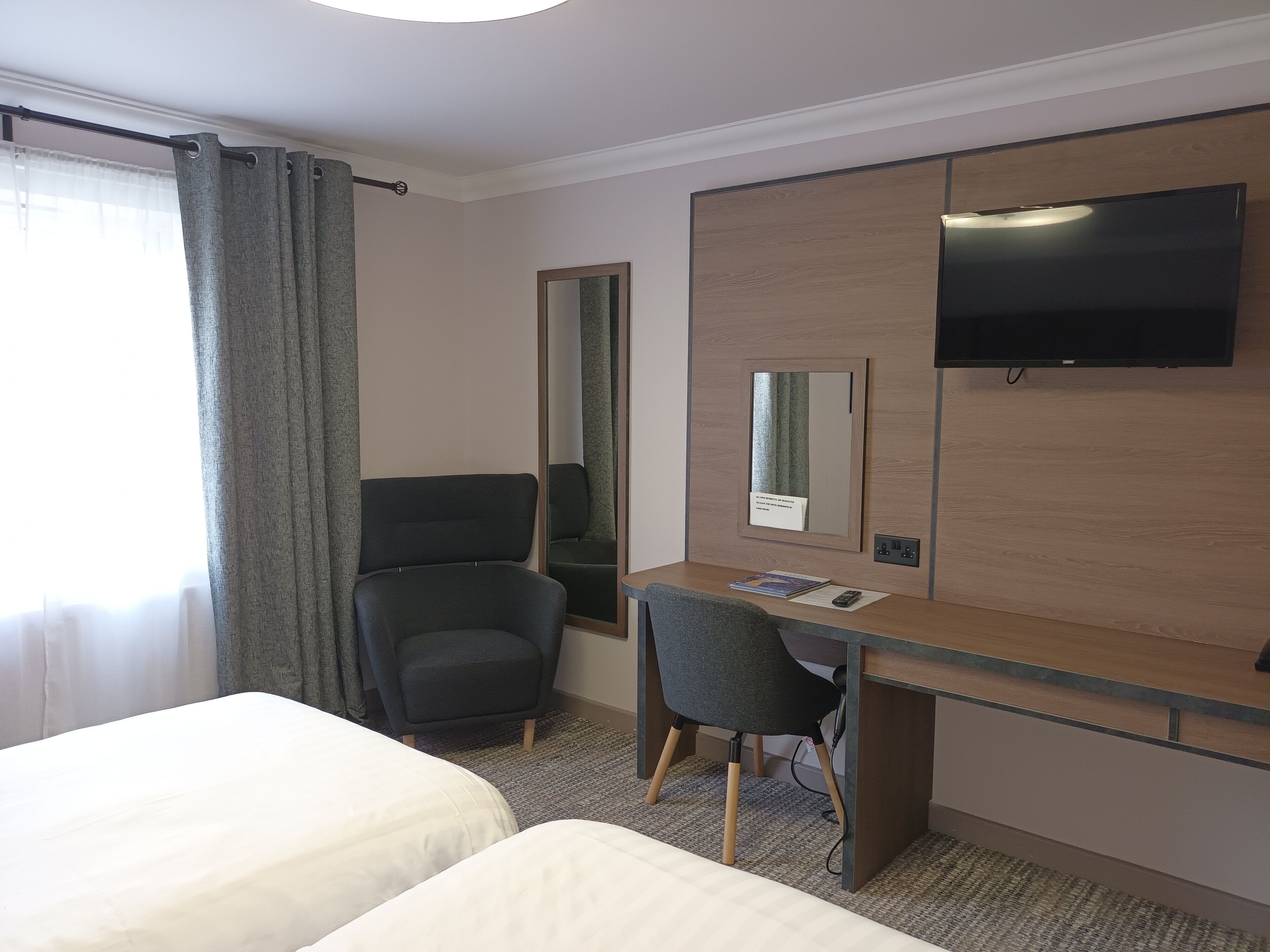A hotel room with two beds, a desk with a chair, a flat-screen TV, a mirror, and a window with curtains.