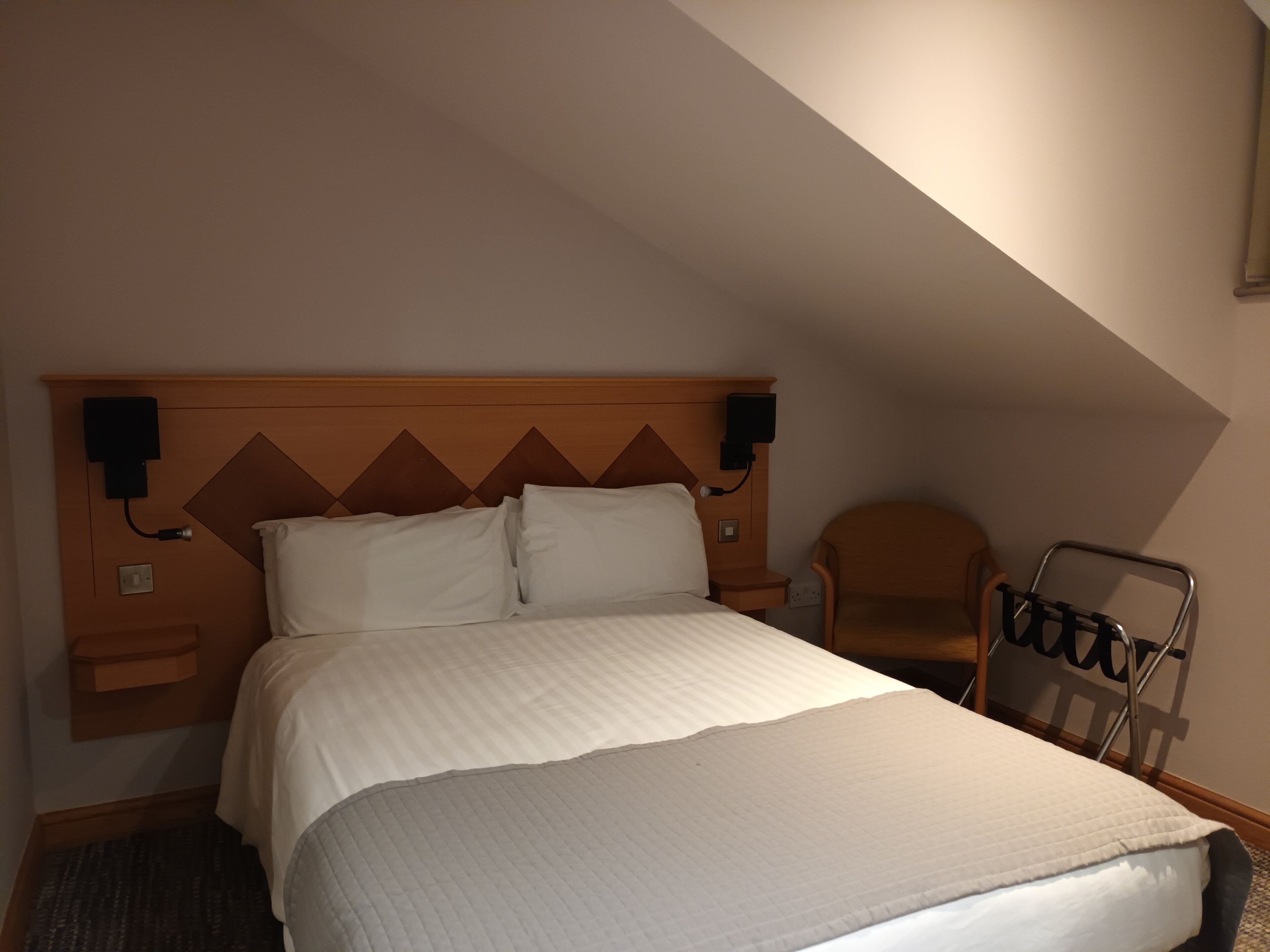 Habitación de hotel con una cama grande, cabecera de madera, dos mesitas de noche con lámparas, una silla y una pequeña mesa con otra silla.