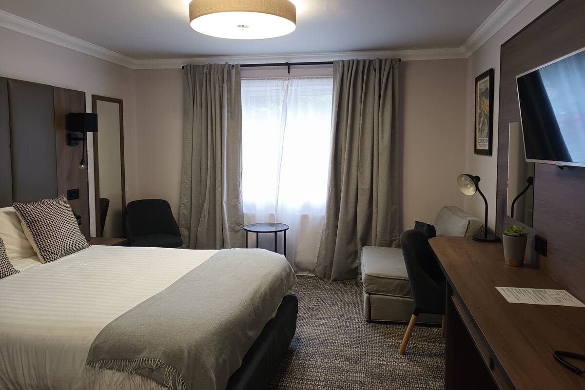 A hotel room with a bed, a desk, a chair, a small table, a lamp, a TV, and a window with curtains.
