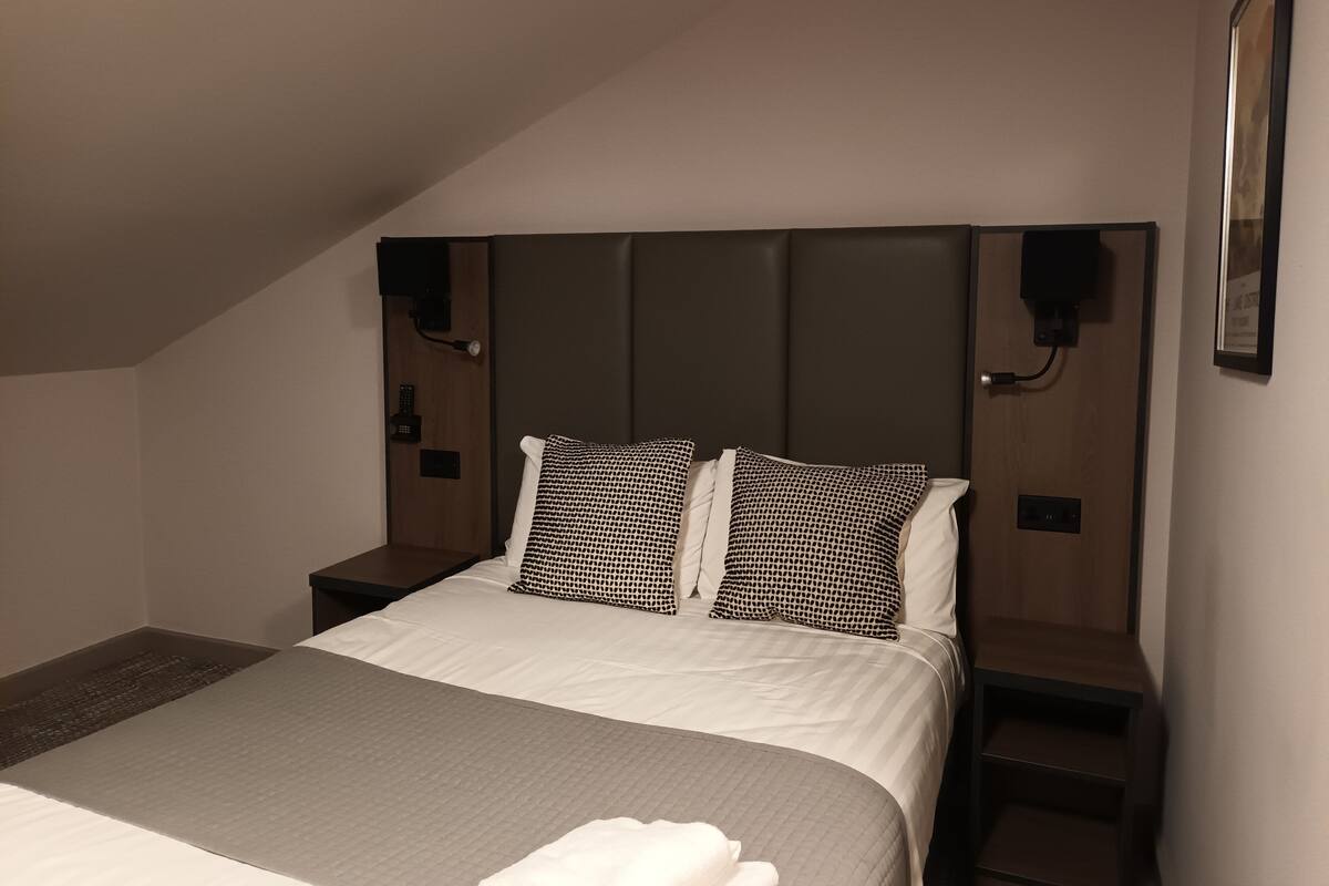 A neatly made bed with a grey blanket and two patterned pillows, a wooden headboard, bedside tables with lamps, and a framed picture on the wall.