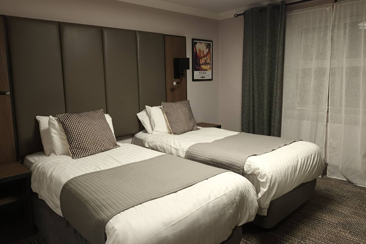 A hotel room with two beds, a headboard, a poster on the wall, and a window with curtains.