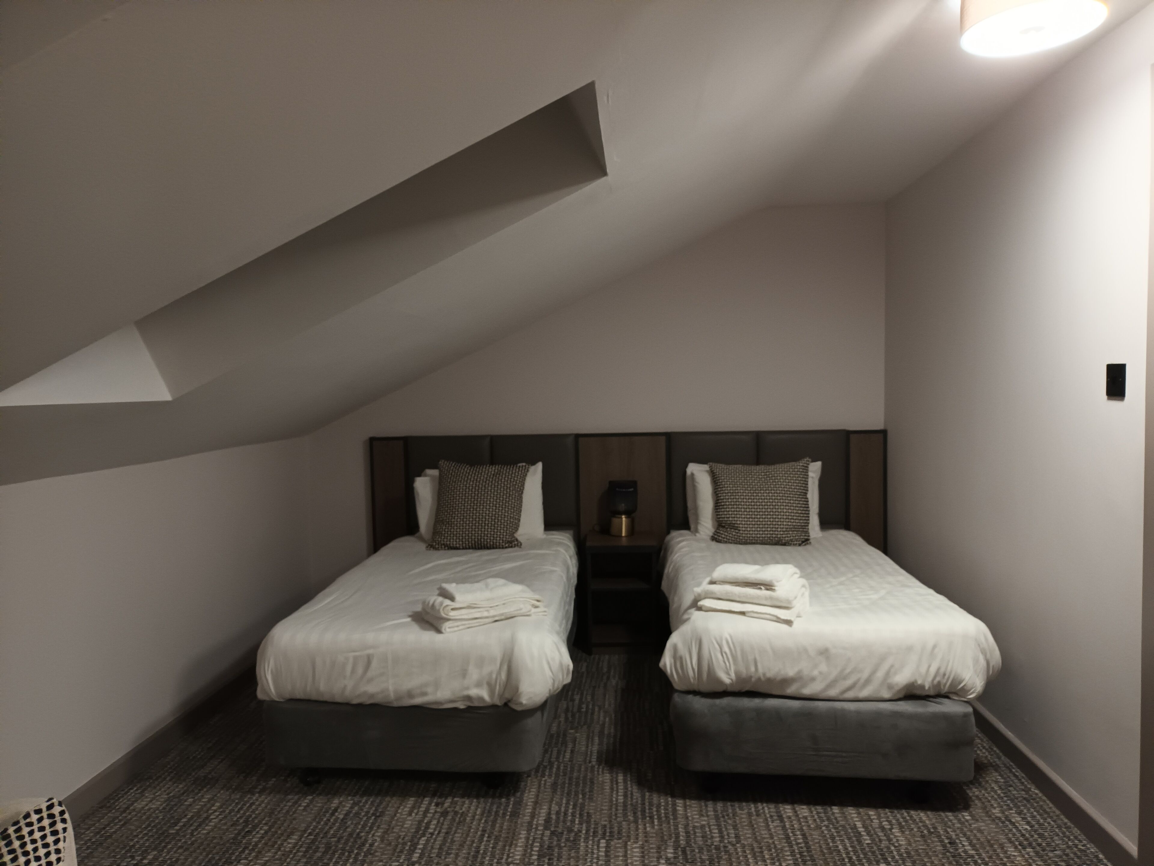 Quarto de hotel moderno com duas camas, uma pequena mesa de cabeceira com abajur e piso coberto por carpete.
