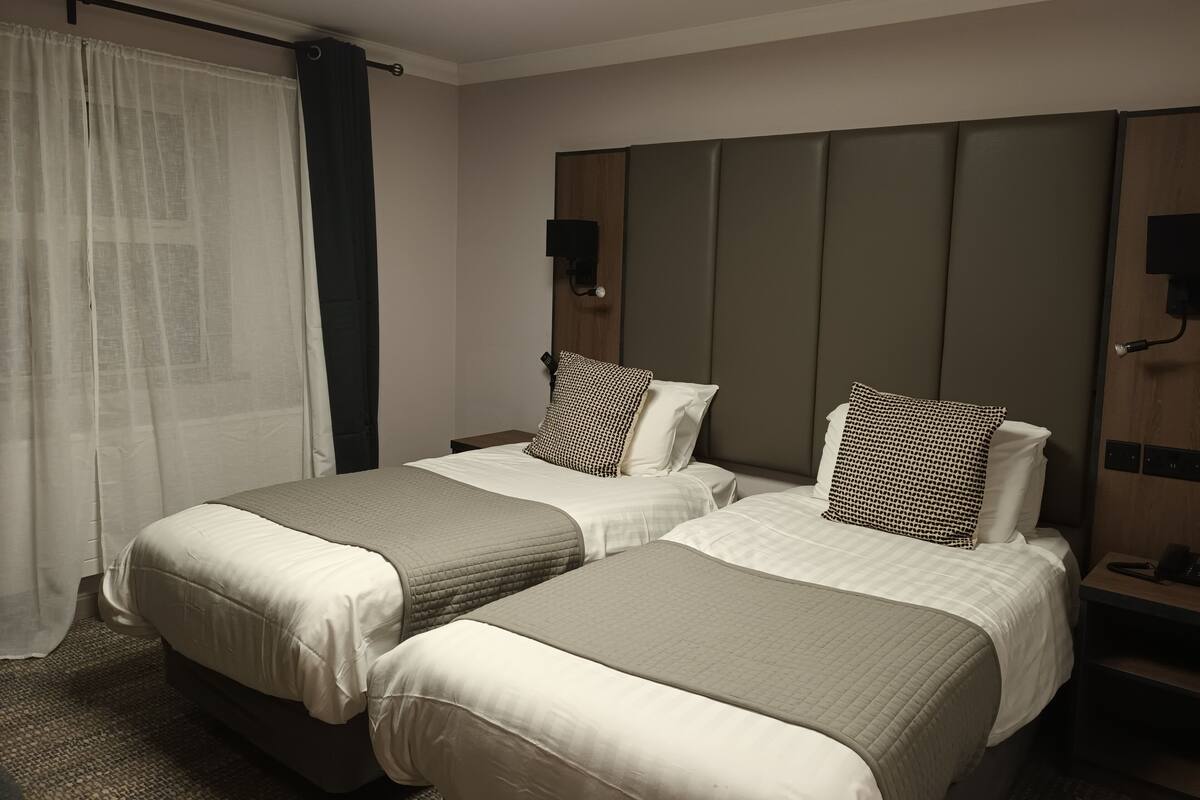 A hotel room with two beds, a large mirror, a bedside table with a lamp, and a window with curtains.