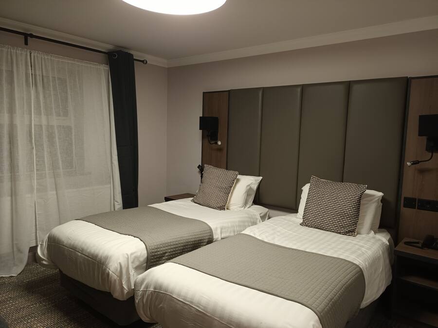 Quarto de hotel com duas camas, um espelho grande, mesa de cabeceira com abajur e janela com cortinas.