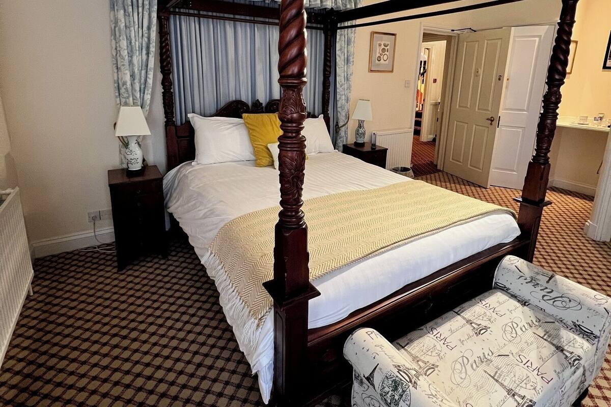 A four-poster bed with a checkered floor, a bedside table with a lamp, and a chair with a Paris-themed cover.