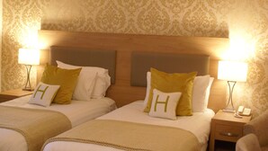 A hotel room with two beds, each with a headboard, a bedside table with a lamp, and a nightstand with a phone. - Haley's Hotel (Leeds)