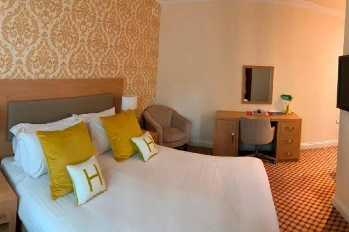 A hotel room with a large bed, bedside tables, a desk, a chair, a TV, and patterned wallpaper.